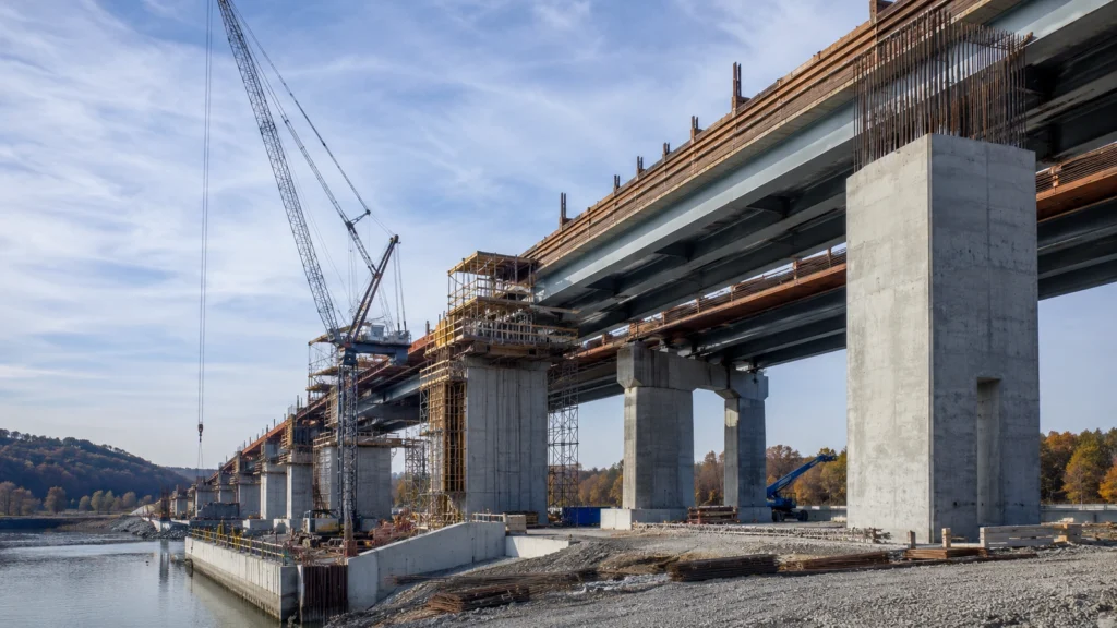 Bridge and transportation structure under construction with concrete piers, steel girders, formwork, rebar, scaffolding, and crane operations.