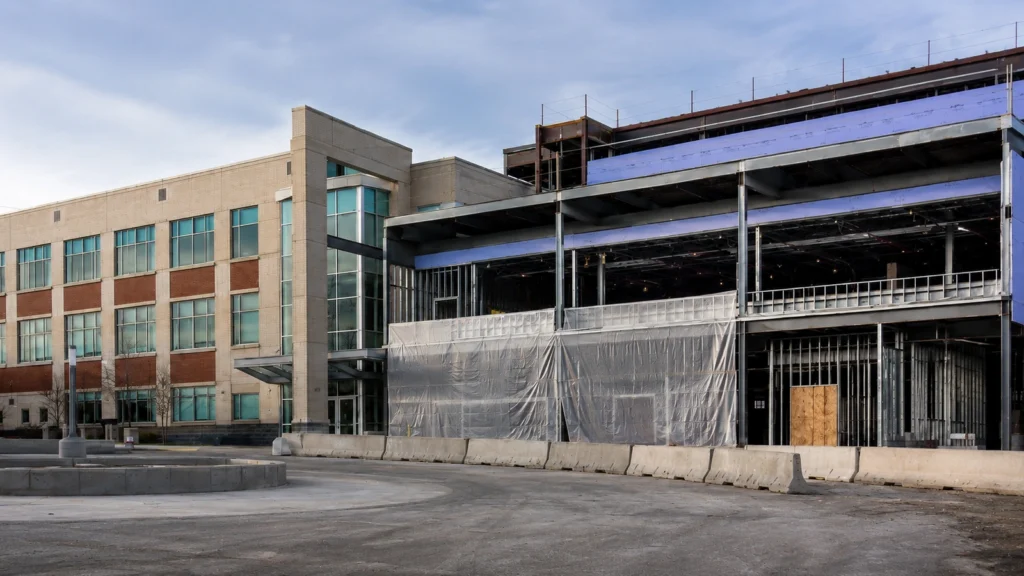 Institutional building undergoing renovation and expansion with existing facade, new steel framing, and temporary protection.