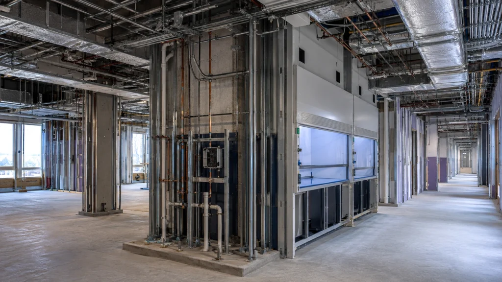 Laboratory and research facility interior under construction with fume hood, ductwork, piping, framing, and utility rough-ins.